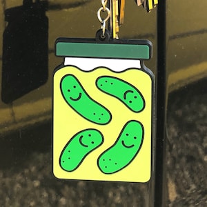 May include: A keychain charm shaped like a glass jar filled with four green pickles. The pickles have a cartoon-style smiley face. The keychain is attached to a silver chain and a set of keys.