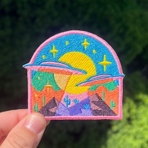 May include: An embroidered patch featuring a desert landscape with two flying saucers, a yellow sun, and stars. The patch is pink, blue, yellow, and green.