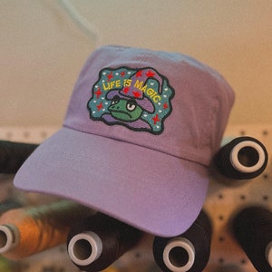 以下が含まれることがあります： ラベンダー色の野球帽。緑色のカエルが魔法使いの帽子をかぶった刺繍パッチが付いています。パッチには「LIFE IS MAGIC」と黄と白の文字で書かれており、赤い星と青い縁取りで囲まれています。