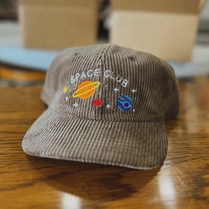 以下が含まれることがあります： 惑星と星が刺繍された茶色のコーデュロイ製のベースボールキャップ。キャップの前面には「SPACE CLUB」という文字が刺繍されています。