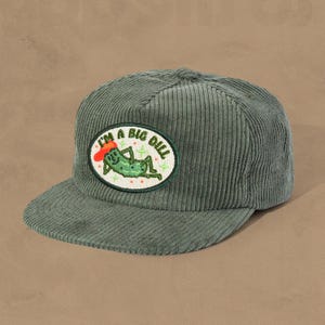 May include: A sage green corduroy baseball cap with a flat peak. The cap has an embroidered patch with the text "I'M A BIG DILL" and a cartoon pickle wearing a red hat. A casual accessory.