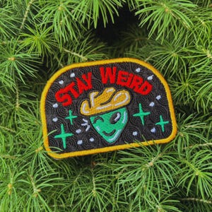 May include: A black embroidered patch with yellow stitching. The patch features a green alien wearing a yellow cowboy hat and the text "STAY WEIRD" in red.