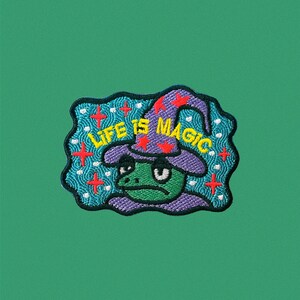 May include: A green frog wearing a purple hat with red stars and the text "LIFE IS MAGIC" in yellow letters. The frog is surrounded by a blue and white background with red crosses.
