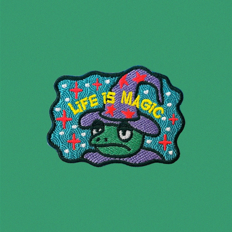 May include: A green frog wearing a purple hat with red stars and the text "LIFE IS MAGIC" in yellow letters. The frog is surrounded by a blue and white background with red crosses.