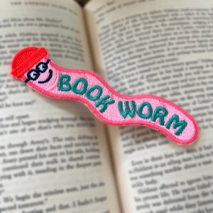 Puede incluir: Marcapáginas de gusano de libro rosa con un sombrero rojo y ojos saltones. El marcapáginas está bordado con el texto "BOOK WORM" en verde.