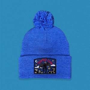 May include: A blue knit beanie with a pom pom on top and an embroidered patch that says "Cryptid Club" with a black silhouette of a creature flying over a cityscape.