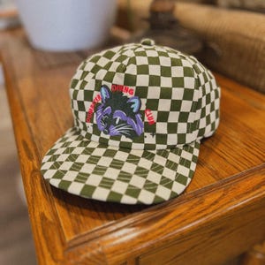 May include: A green and white checkered baseball cap with an embroidered raccoon and the text "Dumpster Diving Club".