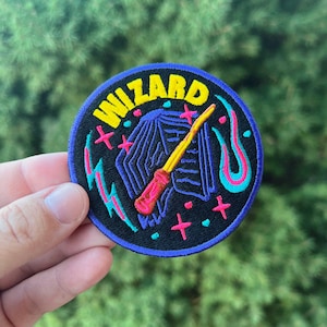 May include: A blue and black embroidered patch with the word "WIZARD" in yellow letters. The patch features a purple and blue wizard's hat with a pink and yellow wand.