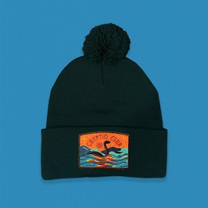 May include: A dark green knitted beanie with a pom pom on top. The beanie has an embroidered patch on the front that says "Crypto Club" and features a cartoon monster in the water.