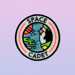 May include: A white embroidered patch with a red and white circle around a blue and green globe with a black silhouette of a person's head. The text "SPACE CADET" is written in black around the globe.