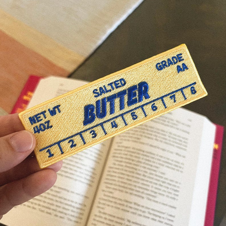 May include: A yellow and blue embroidered patch that reads "SALTED BUTTER" with a scale of numbers from 1 to 8 and "NET WT 4OZ" and "GRADE AA".