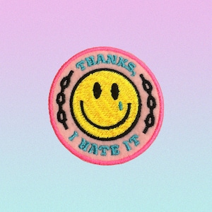 Puede incluir: Parche bordado rosa con una cara sonriente amarilla con una lágrima azul, rodeado de un diseño de eslabón de cadena. El texto "Gracias, lo odio" está escrito alrededor de la cara sonriente.