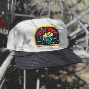 May include: White and black baseball cap with a green alien wearing a cowboy hat and the text "Stay Weird" on a patch.