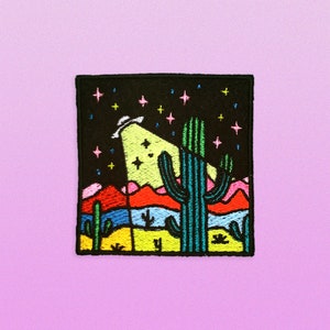 May include: Embroidered patch featuring a desert landscape with a large cactus in the center, a UFO in the sky, and a starry night background. The colors are bright and vibrant, including red, blue, yellow, green, and pink.