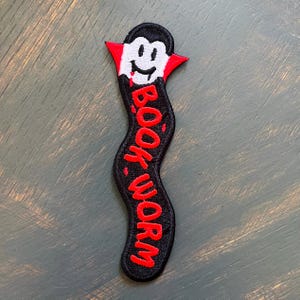 Puede incluir: Parche bordado negro y rojo con forma de gusano. La parte superior del parche presenta una cara de vampiro sonriente con un cuello rojo. Las palabras "BOOK WORM" están bordadas en rojo en el cuerpo del gusano.