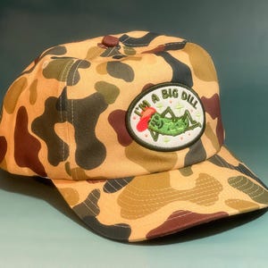 Puede incluir: Gorra de béisbol de camuflaje con visera y botón marrón. La gorra presenta un parche bordado con el texto "I'M A BIG DILL" y un personaje de dibujos animados de pepinillo.