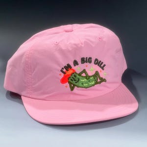 Puede incluir: Gorra de béisbol rosa con visera curva. La gorra presenta un diseño bordado de un pepinillo de dibujos animados con el texto "I'M A BIG DILL". Hecha de un material ligero.