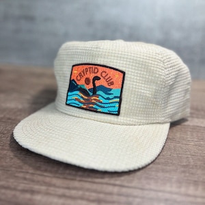 May include: A white corduroy baseball cap with a black embroidered patch that says "Cryptid Club" and features a cartoon image of a Loch Ness Monster.