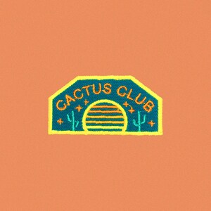May include: A blue and yellow embroidered patch with the text "Cactus Club" and a stylized sunset graphic.