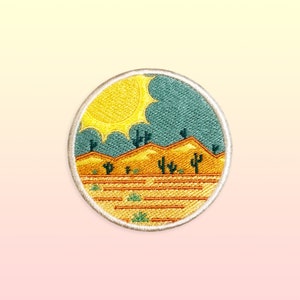 May include: A round embroidered patch with a desert scene. The patch features a yellow sun, green cacti, and brown mountains against a blue sky. The patch is surrounded by a white border.