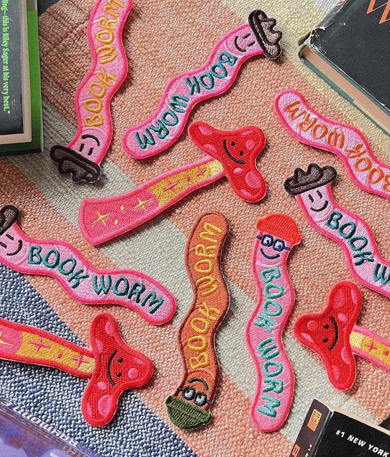 K&ouml;nnte beinhalten: Ein Set aus neun bunten, gestickten B&uuml;geletiketten mit einem Wurm und dem Text "Book Worm". Die Etiketten sind in verschiedenen Farben, darunter Rosa, Orange und Gr&uuml;n. Einige Etiketten zeigen einen Pilz oder eine Brille auf dem Wurm.