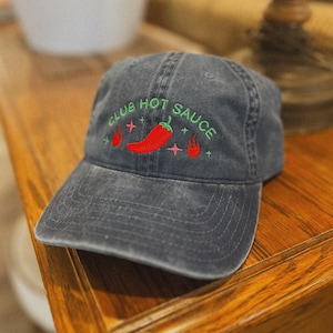 May include: A grey baseball cap with the embroidered text "CLUB HOT SAUCE" in green. A red chilli pepper and flames are embroidered below the text. The cap is on a wooden surface.