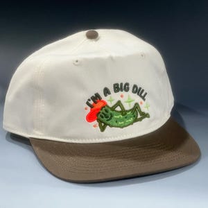 Puede incluir: Gorra de béisbol blanca con visera marrón. La gorra presenta un bordado de un pepinillo verde con un sombrero rojo, con el texto "I'M A BIG DILL" en letras negras.
