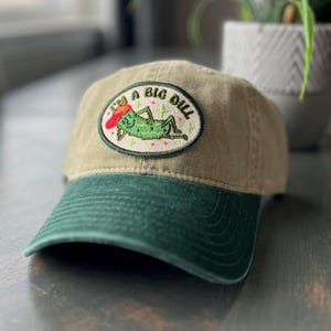 May include: A green and brown baseball cap with a green embroidered patch that says "I'm a Big Dill" and features a cartoon pickle wearing a red hat.