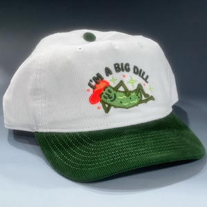 May include: White corduroy baseball cap with a green brim. The cap features an embroidered design of a pickle with the text "I'M A BIG DILL" in green, red, and gold. The cap has a button on top and is a casual accessory.