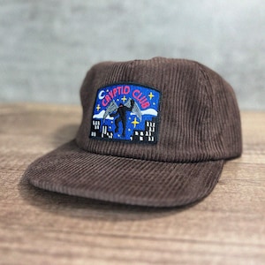 May include: A brown corduroy baseball cap with a patch featuring a black silhouette of a creature with wings, the text "CRYPTID CLUB", and a blue background.