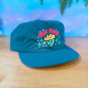 May include: Teal baseball cap with a curved peak. The cap has the embroidered phrase "Stay Weird" above an alien wearing a cowboy hat. The alien is green with a yellow hat and a smiling face. The background is a blurred blue and green.