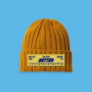 May include: Mustard yellow knit beanie with a rectangular label that reads "SALTED BUTTER" in blue text. The label also includes "NET WT 4OZ" and "GRADE AA". The beanie is set against a light blue background.