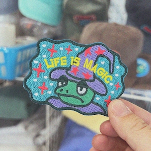 May include: A blue and purple embroidered patch featuring a frog wearing a wizard hat. The patch reads "Life is Magic" in yellow lettering.