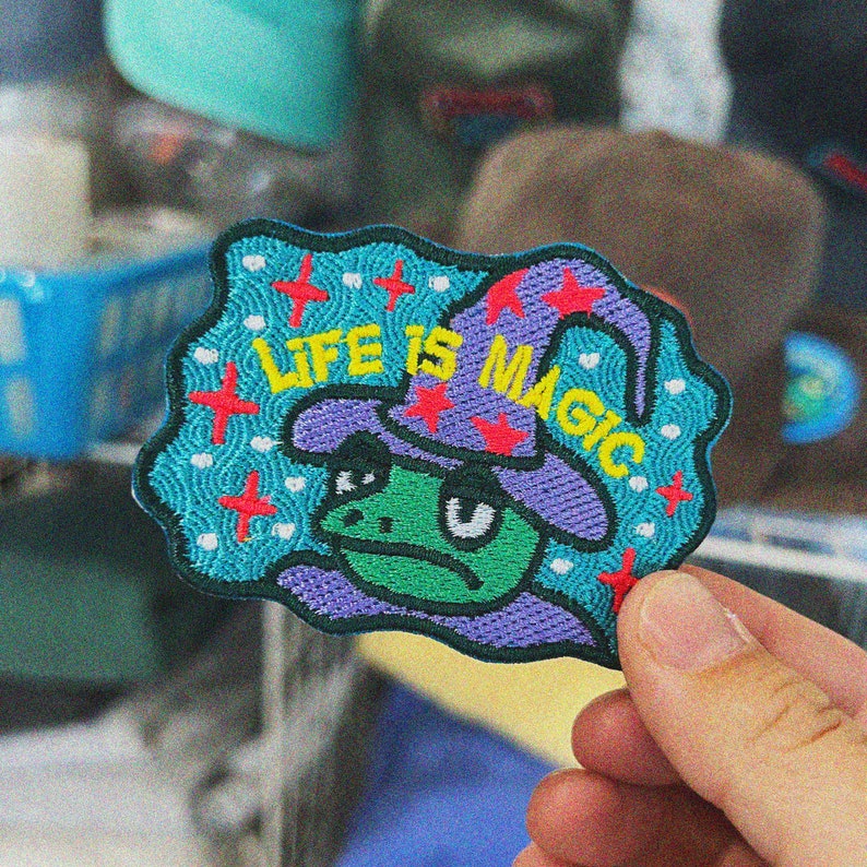 May include: A blue and purple embroidered patch with a frog wearing a wizard hat. The patch says "Life is Magic" in yellow letters.