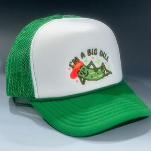 May include: A green and white trucker hat with the text "I'M A BIG DILL" embroidered on the front. The hat features a green brim and mesh back, with a white front panel displaying a cartoon dill pickle design.