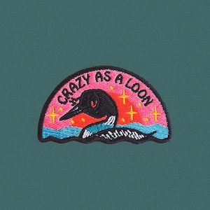 May include: Embroidered patch with a loon design and the text "Crazy as a Loon." The patch features a black loon with a pink and orange sunset background, blue water, and yellow stars. The patch is on a teal green background.