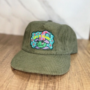 以下が含まれることがあります： 緑色のコーデュロイ製のベースボールキャップ。刺繍されたパッチには、魔法使いの帽子をかぶったカエルと「Life is Magic」の文字がデザインされています。