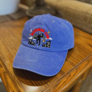 May include: A blue baseball cap with an embroidered design of a creature with wings and the text "Mothman Club".