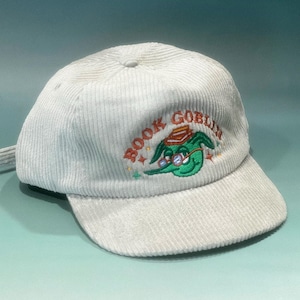May include: A light grey corduroy baseball cap. It features the embroidered text "BOOK GOBLIN" and a green goblin graphic wearing glasses. The cap has a curved peak and an adjustable strap.