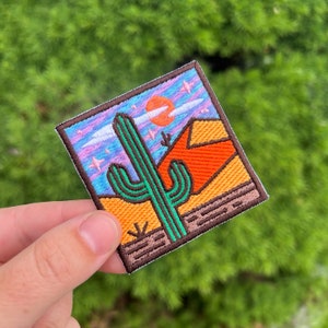Puede incluir: Un parche bordado con una escena del desierto. El parche presenta un cactus verde, una montaña roja y naranja, y un cielo azul con nubes blancas y estrellas. El parche está rodeado por un borde marrón.