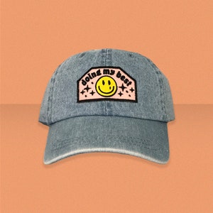 May include: A blue denim baseball cap with a pink patch that says "doing my best" with a yellow smiley face in the centre.