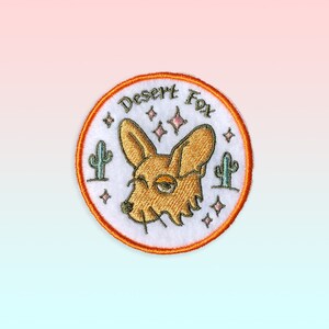 May include: A white, round embroidered patch with an orange border. The patch features a cartoon desert fox with a light brown body and a light green cactus on either side. The text "Desert Fox" is embroidered in green above the fox.