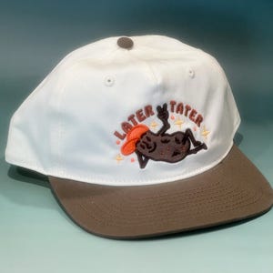 May include: A white baseball cap with a brown brim and a dark brown button on top. The front features an embroidered graphic of a brown potato wearing an orange hat, reclining with a peace sign, and the text LATER TATER in brown letters.