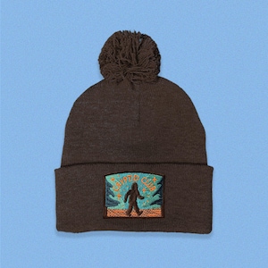 May include: Brown knitted beanie with a pom pom on top and an embroidered patch that says "Crypto Club" with a cartoon Bigfoot walking in front of a blue and orange mountain range.
