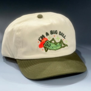 Puede incluir: Gorra de béisbol color crema con visera verde. La gorra presenta un diseño bordado de un pepinillo verde con un sombrero rojo y el texto "I'M A BIG DILL" en verde y rojo.