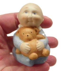 以下が含まれることがあります： テディベアを抱いた赤ちゃんの陶器製フィギュア。赤ちゃんは水色の服を着ており、頭は禿げています。テディベアは茶色です。白い背景を背景に、手でフィギュアが持たれています。