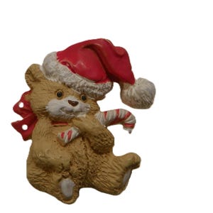 May include: A tan teddy bear figurine wearing a red Santa hat with white trim. The bear has a white muzzle, dark eyes, and is holding a red and white striped candy cane. A red bow is visible behind the bear's head, and the figure has a textured surface.