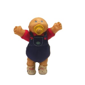 May include: A vintage Cabbage Patch Kids doll. The doll has a tan skin tone, a dummy, and is wearing a red shirt, blue dungarees, and floral-patterned socks. The doll's arms are raised.