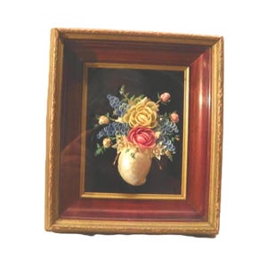 Peut inclure: Une décoration murale encadrée présentant un arrangement floral en trois dimensions composé de coquillages. Le bouquet comprend des fleurs en coquillages jaunes, roses et bleues dans un vase en coquillage blanc sur fond noir.