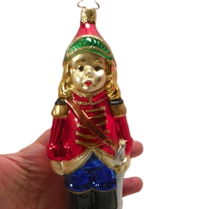 May include: A glass Christmas ornament shaped like a soldier in a red uniform with gold trim, blue trousers, and black boots. The figure has blonde hair, a red hat with a green brim, and a silver sword at the side, topped with a star-shaped wire loop.